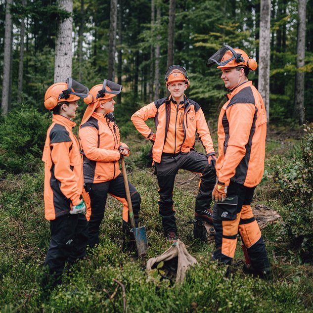 Eine Forstwirtin und drei Forstwirte stehen in Arbeitskleidung und mit einer Schaufel im Kreis auf einer Lichtung im Wald.